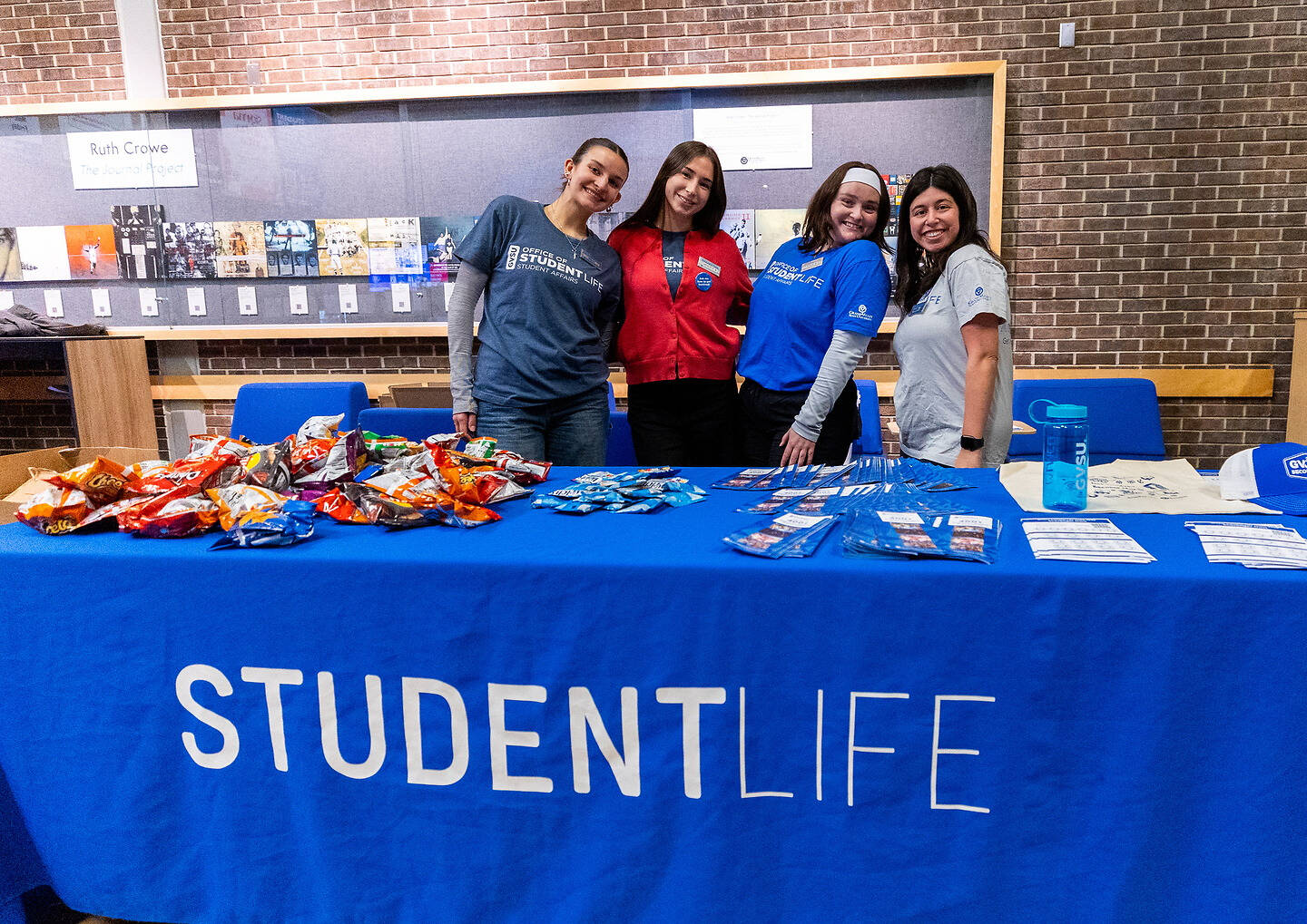 Four individuals stand behind a blue table covered with snacks and promotional materials, smiling for the camera at a StudentLife event.